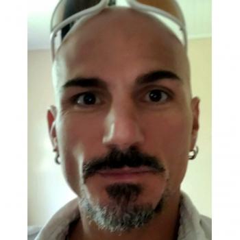 Un homme de type européen, entre 35 et 40 ans, chauve, les yeux marrons, avec une moustache et un bouc poivre et sel, portant une chemise blanche, une paire de lunettes de soleil sur la tête, et des boucles aux deux oreilles.